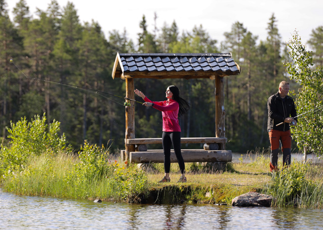 A woman casts her fishing rod at a calm lake while a man stands beside her.
