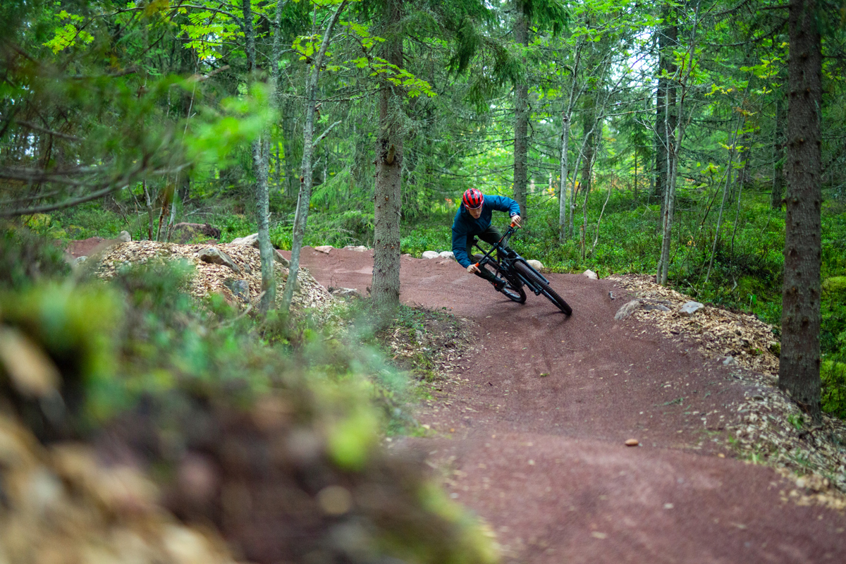 A cyclist wearing a helmet rides through a curve on a red gravel mountain bike trail in a dense green forest.