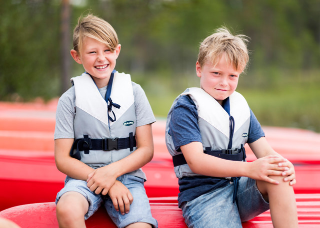 Two smiling children wearing life jackets sit on canoes on a summer day.