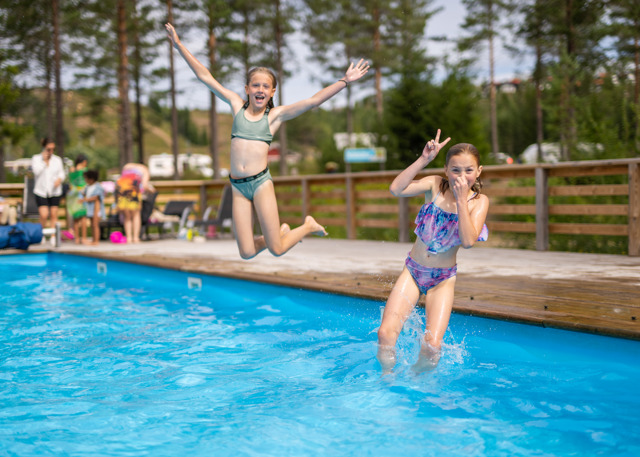 Two children in swimwear happily jump into an outdoor pool on a sunny day.