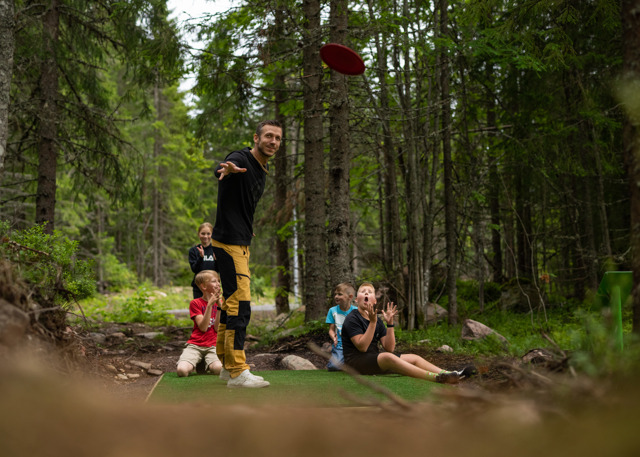 A man throws a frisbee while children and adults watch at a disc golf course in the forest.