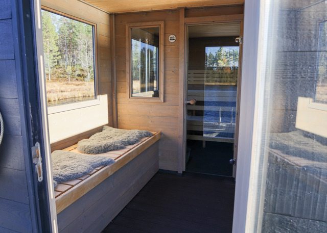 Inside a sauna cabin with windows facing nature and a lake. Wooden interior, benches with sheepskin, and a view over the water.