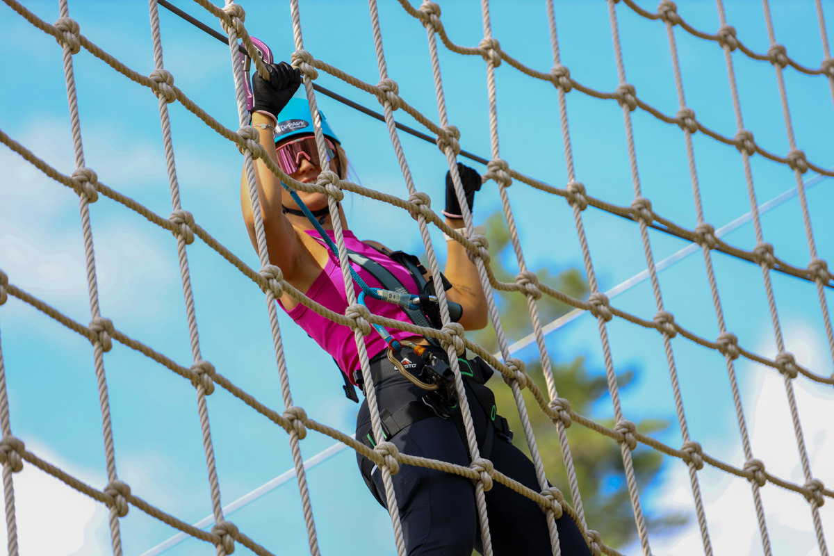 A woman with a safety harness climbs a rope net in a high-altitude course and smiles at the camera.